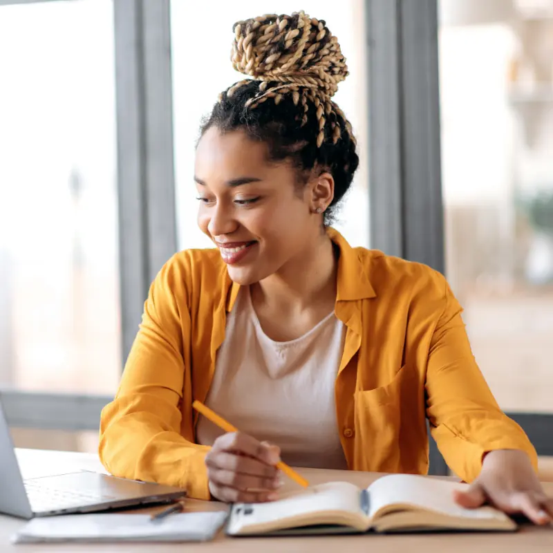 Enseignement en ligne, e-learning. Jeune femme afro-américaine rayonnante, vêtue d'une tenue décontractée et élégante, qui suit des cours à distance sur son ordinateur portable, écoute un cours en ligne et prend des notes chez elle, tout en souriant