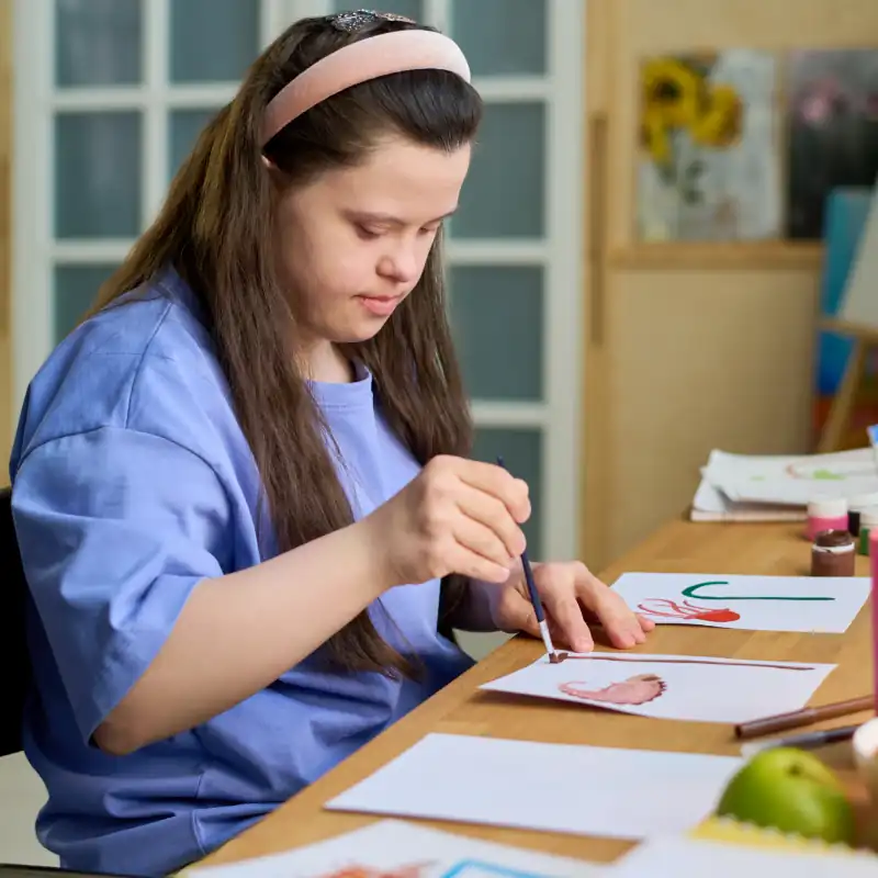 Une jeune fille atteinte d'un handicap mental peint à l'aquarelle avec un pinceau sur du papier, assise à un bureau devant un ordinateur portable dans le salon 